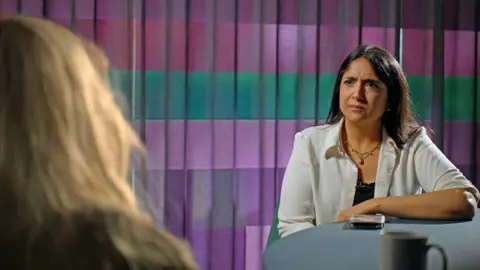 Sima Kotecha, who is wearing a necklace and a white shirt, interviews a woman in a room with colourful curtains at the BBC.