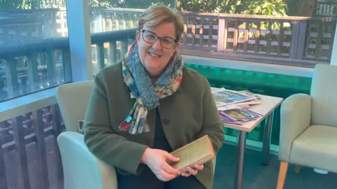 Jane Thompson A woman, with glasses, short hair and wearing a scarf and green jacket, seated and holding a book 