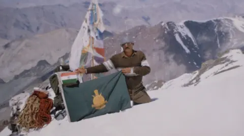 Family handout A man wearing protective clothing on a snow-capped mountain peak holds a green flag, with a smaller Indian flag above it.   