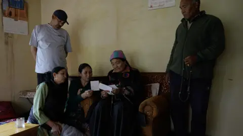 Family members of Morup gathered in a room. His wife is sitting in a chair and looking at old photos.