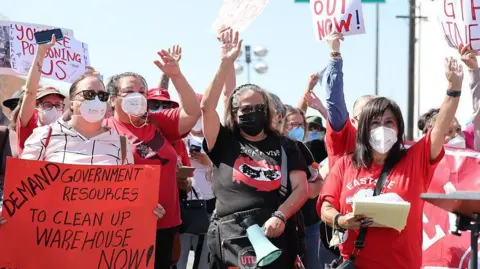 Getty Images Residents in red and white and black shirts carry signs and wear masks and raise their hands during a protest. One sign reads: "Demand government resources to clean up warehouse now!"