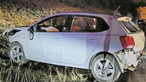 South African police A small white car - damage visible at both ends of the vehicle - 
on a grass verge