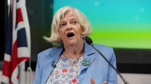 PA Media Ann Widdecombe is wearing a light blue blazer with a floral shirt and a turquoise brooch. Her hair is short and she is speaking in front of a microphone. The background behind her is blurred but a Union Jack can be seen.