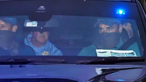 PA Media A close up image looking through the front windscreen of a vehicle. The two men in the front passenger seats are wearing baseball caps and have the bottom half of their face covered. There is a man on the left rear passenger seat wearing a blue shirt with a Gardai emblem. His face is obscured by the rear view mirror. On the right the face of Daniel Kinahan can be seen over the shoulder of one of the front passengers. He has stubble and is looking out the window.
