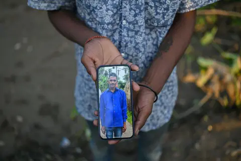 Getty Images Amit Giri, 20, holds a mobile phone displaying a photograph of his father, Siv Sankar Giri, who died in the floods, outside his flood damaged home on August 05, 2026 in Sivasagar, Assam, India.