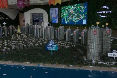 BBC/ Gavin Butler A diorama of a city, with fake ocean in the background, large picket sign saying "land to be developed" in a fake green space of trees, and model high-rise buildings behind that, some of them with signs saying "fast selling". On the wall behind the diorama is a large screen showing a map, and a logo saying "Forest City".