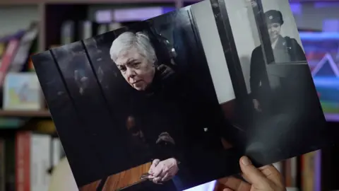 A hand holds a printed out photograph of Nadezhda Buyanova, a 68-year-old Ukrainian-born paediatrician in the dock at court, guarded. File photo