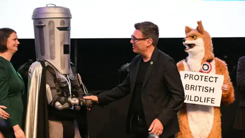 Getty Images Burnham is in the centre of the photograph, wearing a black suit over a black t-shirt, and shaking hands with Count Binface on his left. To his right is a man dressed as a fox, holding a placard reading Protect British Wildlife.