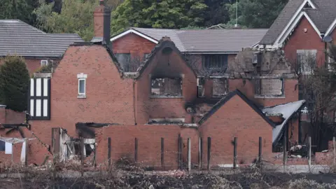 EPA A red brick house left in ruins from fire, with its roof burned off, and black singes around the edges of the ruins. Burnt shrubbery can be seen in front of it, with houses behind still in tact.
