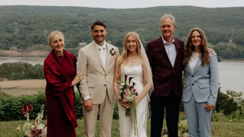Jennifer Morris photography Michele Heisler stands next to her son on his wedding day, flanked by her husband and daughter. She wears a red velvet shawl and dress, while her son wears a beige suit.