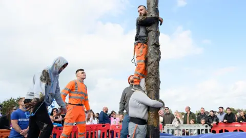 Gregor Campbell Photography A group of men attempt the greasy pole challenge - one man is standing on another's shoulders, with his arms wrapped around the pole. Another four men are watching, presumably ready to start climbing.