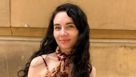 A picture of a young woman with black hair in a floral dress smiling slightly in front of a beige brick building.
