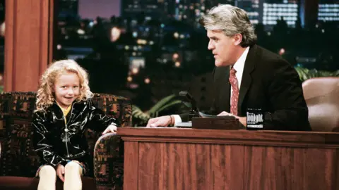 Getty Images Actress Hayden Panettiere, who has curly blonde hair and wears a black shiny jacket, during an interview with host Jay Leno on October 3, 1996 