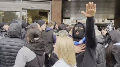 Crowd gathered outside a building entrance, with several people in dark hooded jackets and one individual raising an arm while holding a scarf around the neck, others recording with smartphones.