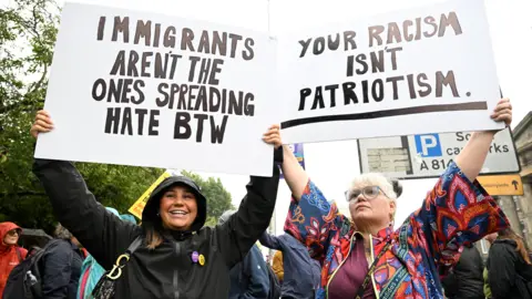 PA Media People at a public demonstration hold large placards above their heads. One sign reads, “IMMIGRANTS AREN’T THE ONES SPREADING HATE BTW,” and another reads, “YOUR RACISM ISN’T PATRIOTISM.” Other demonstrators, trees, and road signs are visible in the background.