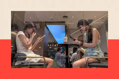 Getty Images Two young women sit at a table on their phones