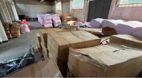 Large numbers of boxes inside a warehouse - alongside dinghies and lifejackets  in plastic bags