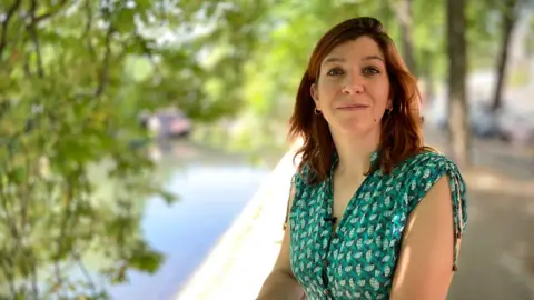 Marie-Hélène Brice is wearing a green patterned, short-sleeved dress. She has straight, shoulder-length brown hair. Behind her are some trees and a stretch of water.