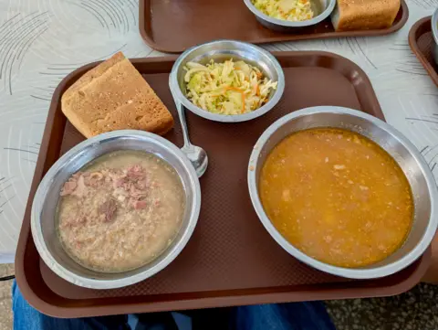 Three metal bowls on a tray with a hunk of bread. Two are soups, one with meat and there is a salad in the third bowl.