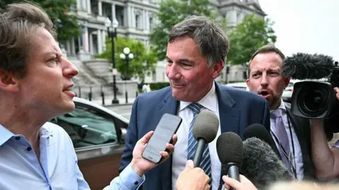 AFP via Getty Images LeBlanc is pictured surrounded by reporters on a side walk in Washington, DC. He has short black and grey hair and is listening to a reporter asking him a question. The reporter holds up an iPhone to LeBlanc's face to record him. Others hold up microphones to his face. A TV camera is on his right. He is wearing a navy blue suit and a striped blue tie.