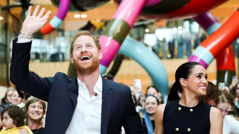 Reuters On the left, Prince Harry smiles, looks up and waves his right hand in the air, and  on the right, Meghan smiles and looks to her left in front of a large crowd and a large colourful scuplture