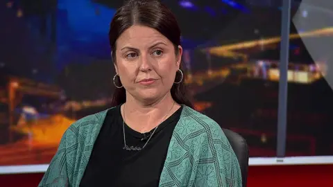 Michaela Rafferty  is wearing a dark top with a green cardigan over it. She has dark hair, is wearing large hoop earrings and a silver necklace.  She is pictured in a television studio.