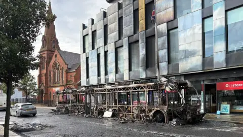 PA Media A Glider bus was set on fire in east Belfast. A burned out bus is in the foreground on a road which has visible marks of a fire. The bus is beside a large pale coloured building with lots of windows, and bearing scorch marks. To the rear of the picture is a red building, with a tall spire.