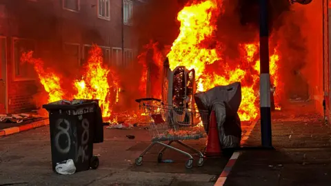 PA Media Bins and a car are set on fire on a street. Huge yellow and red flames are seen coming out of the items with smoke billowing into the air. There are houses to the left of the picture, and more to the right. A lamppost is visible to the right.