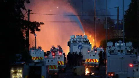 Getty Images Police use water cannon as they block the road from crowds. A large number of police branded armoured vehicles block the road. There are pockets of fires seen between the vehicles and a stream from the water cannon. 