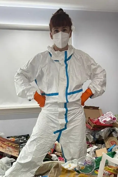 Victoria Gordon A woman standing on a pile of rubbish inside a house. She has ginger hair tied back and a fringe. She is wearing a white hazmat suit, a white face mask and orange gloves.