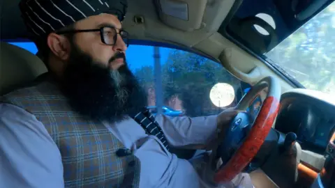 Side shot of a man driving a car, photographed from the passenger seat. He has a full beard, glasses, black headdress with thin white stripes, a light blue top and checked waistcoat.  