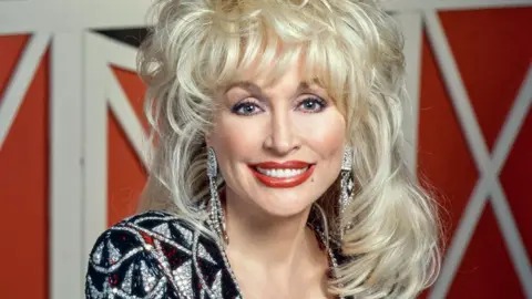 Getty Images/CBS photo Archive A head and shoulders shot.  Dolly's blonde hair stands up in big waves and falls onto her shoulders.  She wears a sliver and black diamante V neck dress with red accent colours.  Long diamante earrings hang down.