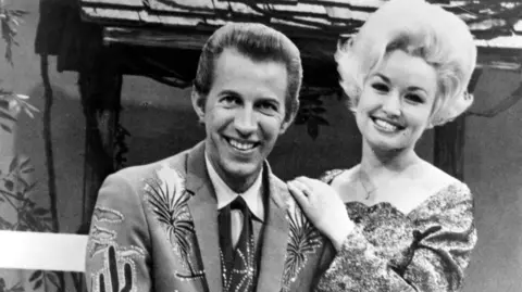 Getty Images/Michael Ochs Archives Wagner sits on a stool in a suit with emblems of cacti and plants in this black and white photogragh. He is posing with his guitar.  Dolly stands behind him leaning on his shoulder.  Her hair is short but still standing on end. 