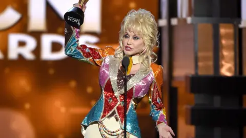 Getty Images/Ethan Miller Dolly holds up the award in the air.  She wears a riding jacket type coloured shiny, sparkly jacket, with cream jodhpurs.