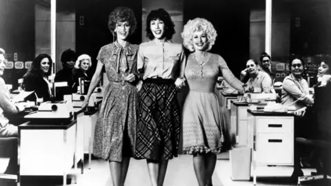 Getty Images/Michael Ochs Archives The three link arms and are pictured walking through the office with desks in the background of this black and white photo.  The female typists in the background are all smiling and looking at the three ladies.