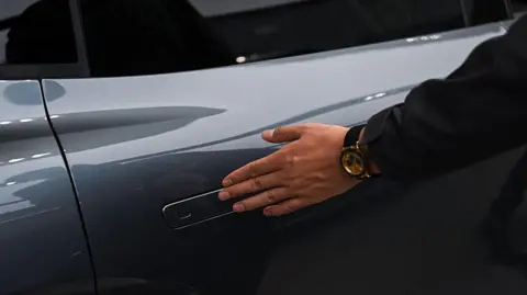 AFP via Getty Images A man inspects an electric car, brushing his hand over its retractable door handle, during a vehicle roadshow in China.