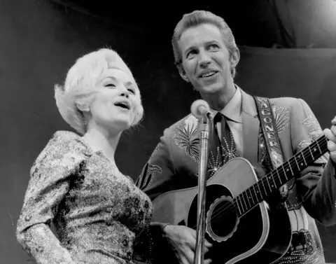 Michael Ochs Archives/Getty Images Black and White image of Dolly and Porter singing into a shared microphone, and he is playing a guitar.