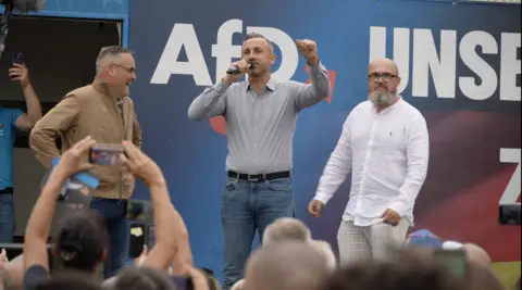 Three men are on-stage at an AfD rally. Ulrich Siegmund is in the centre holding a microphone. Crowds are listening and filming on their phones.