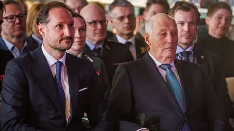 Getty Images Two men in suits sit sombrely listening in an audience - the older man sits on the right