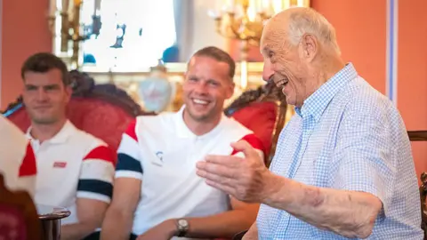 NTB/AFP via Getty Images The king wearing a checkered collared t-shirt laughs as he talks. Two footballers seated on a sofa laugh in the background. 