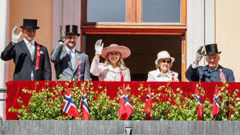Per Ole Hagen/Getty Images Three men and two women dressed in hats and formal wear stand on a balcony waving, flags in front of them.
