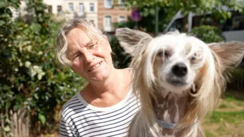 Muriel sits in a green garden wearing a striped white and black t shirt, with her dog Cheva sitting on her lap