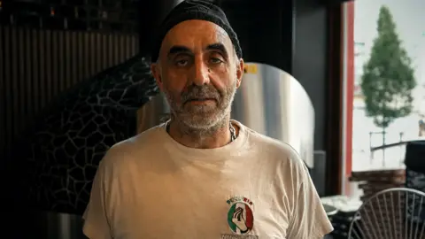 Salvatore stands in a white t shirt and black beanie in his pizzeria in Brussels