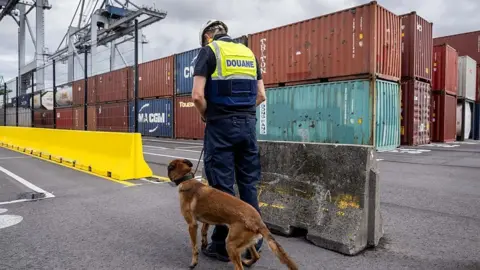 AFP via Getty Images A customs officer and a sniffer dog stand next to containers as they demonstrate their skills during a press tour at the Antwerp port in Doel on July 7, 2025