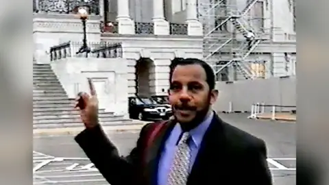 SDNY A screenshot of Bayoumi, wearing a suit with a purple shirt, standing outside the Capitol. He is pointing towards the building.
