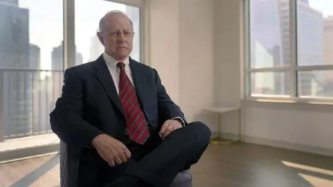 Richard Lambert, who has now retired from the FBI, is interviewed in an office by the BBC. He wears a suit with a white shirt and red tie and has his legs crossed.
