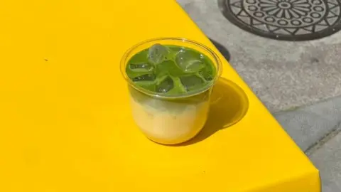 Cantoast Bakery A plastic cup, half filled with a yellowy cream and half filled with bright green matcha and ice cubes, is pictured on a yellow table outside