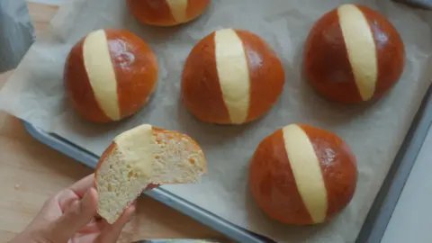 Supreedha Iyer A tray of five shiny brioche buns, cut in the middle and filled with cream, are seen on a sheet of baking paper on a baking tray. A hand is seen holding another bun, cut in half, in the foreground.