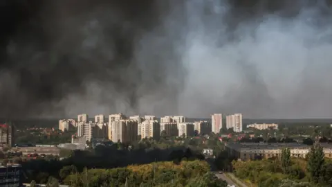 Reuters Black smoke billows into the sky following a strike near a residential area. Several white high-rise buildings feature at the centre of the frame, with a tree-lined road visible in the bottom right