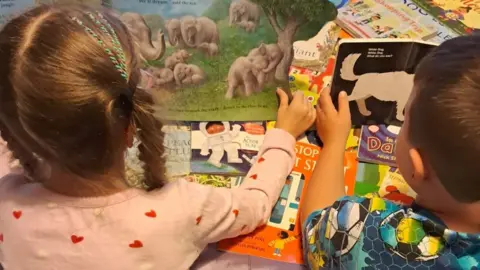 Helen Cashell Two children in pyjamas, their backs turned to the camera, each hold books open - atop a pile of other books they received from Parton's Imagination Library